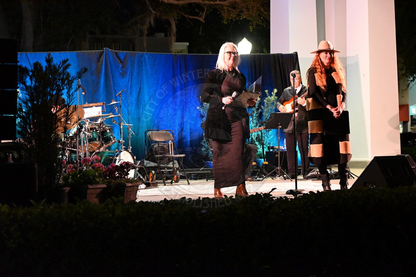 Two women stand on an outdoor stage at night, one holding papers and the other wearing a hat and boots. Behind them, musicians play guitar and drums. Plants and a blue backdrop decorate the stage.