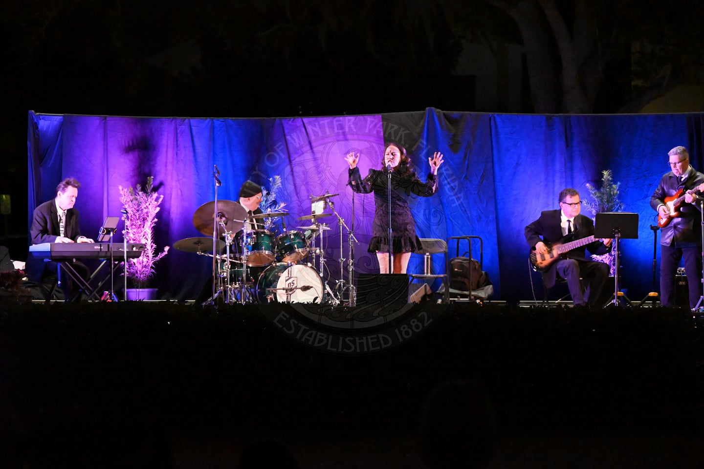A five-member jazz band performs on an outdoor stage at night, with purple-lit curtains behind them. The singer stands center stage, passionately singing, while the other musicians play keyboard, drums, guitar, and bass guitar.
