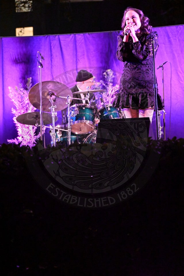A woman in a black dress sings into a microphone on stage, with a drummer playing behind her. The stage is lit with purple lighting and decorated with plants.