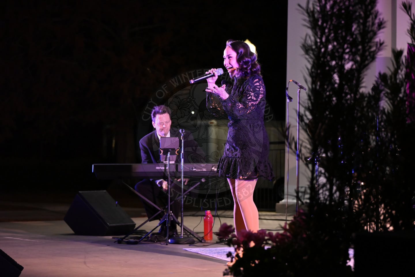 A woman in a black dress sings into a microphone on an outdoor stage at night, while a man plays a keyboard beside her. Trees and stage lights are visible in the background, with plants in the foreground.
