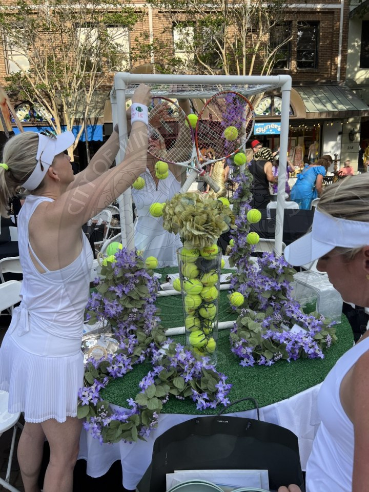 a table decorated with tennis ball things. There is a lady sticking up tennis rackets into a vase filled with tennis balls.