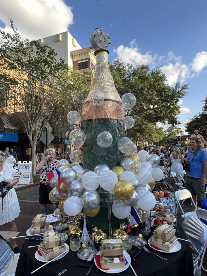 10 foot tall 3 feet wide home made champagne bottel sitting on top of a table with balloons that are intimating bubbles from champagne.