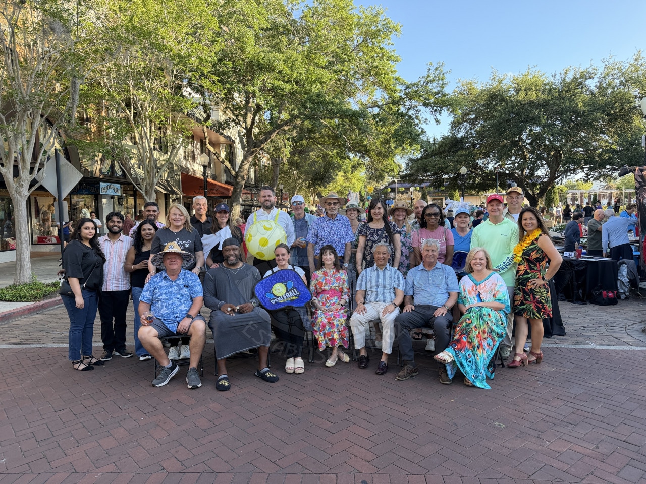 A large group of people pose together outdoors on a brick street, smiling, with trees and buildings in the background.