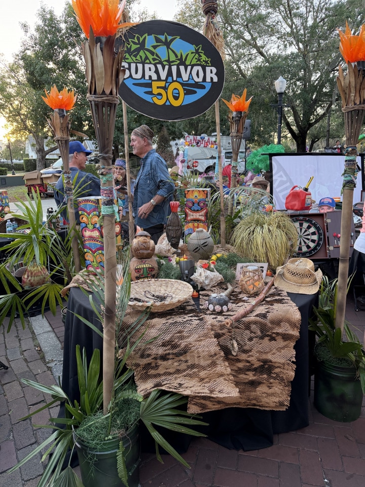 A Survivor 50 themed table with tiki decorations, torches, and tropical plants set up outdoors at an event.