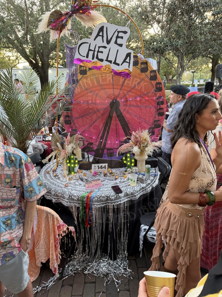 A table decorated with a Ferris wheel backdrop, Ave Chella sign, beads, and festival-themed items; people stand nearby.