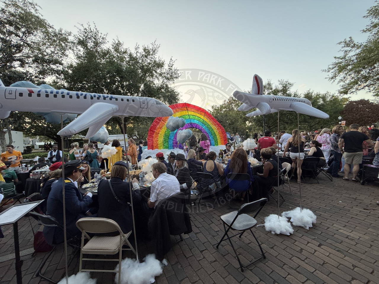 People dining outdoors by rainbow decor, with inflatable airplanes and trees in the background, at a festive event.