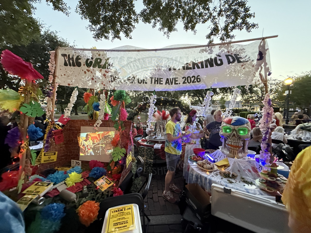 Colorful outdoor event booth with leis, flowers, a skull decoration, and a banner above a crowd under trees at sunset.