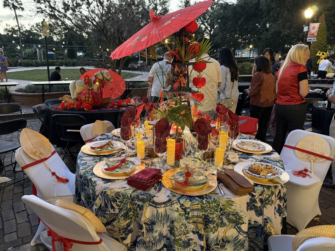 Elegant outdoor table setting with red and gold Asian decor, candles, food, and people gathered around.