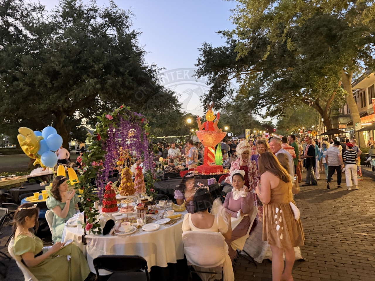 People in costumes sit and stand around decorated tables at an outdoor event with trees and balloons in the background.