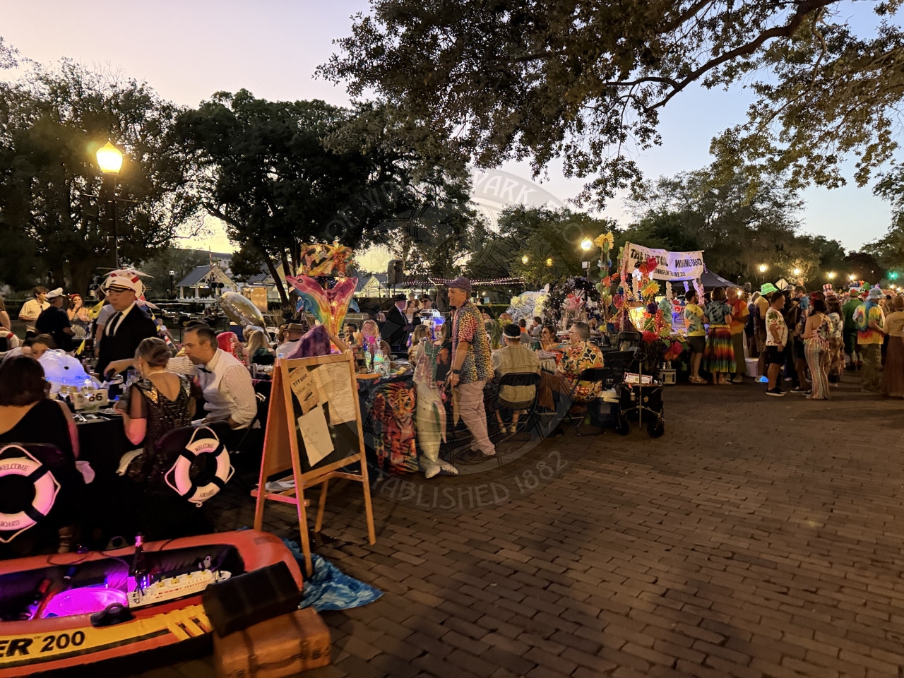People in colorful costumes gather at an outdoor festival with decorations, tables, and trees at sunset.