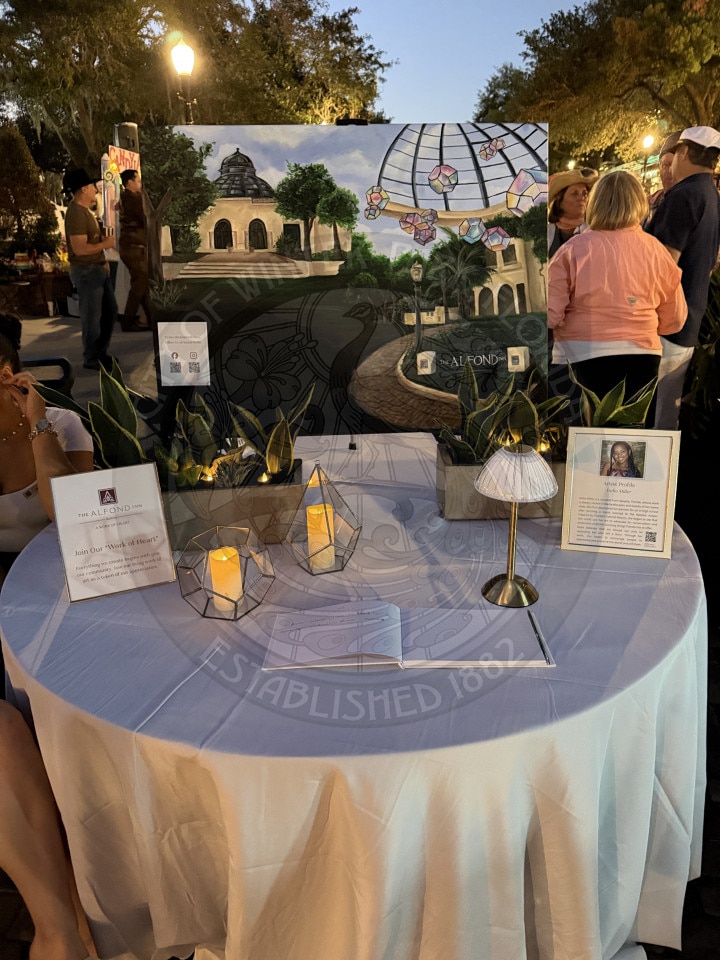 A round table with candles, plants, and artwork on display at an outdoor event with people gathered around.