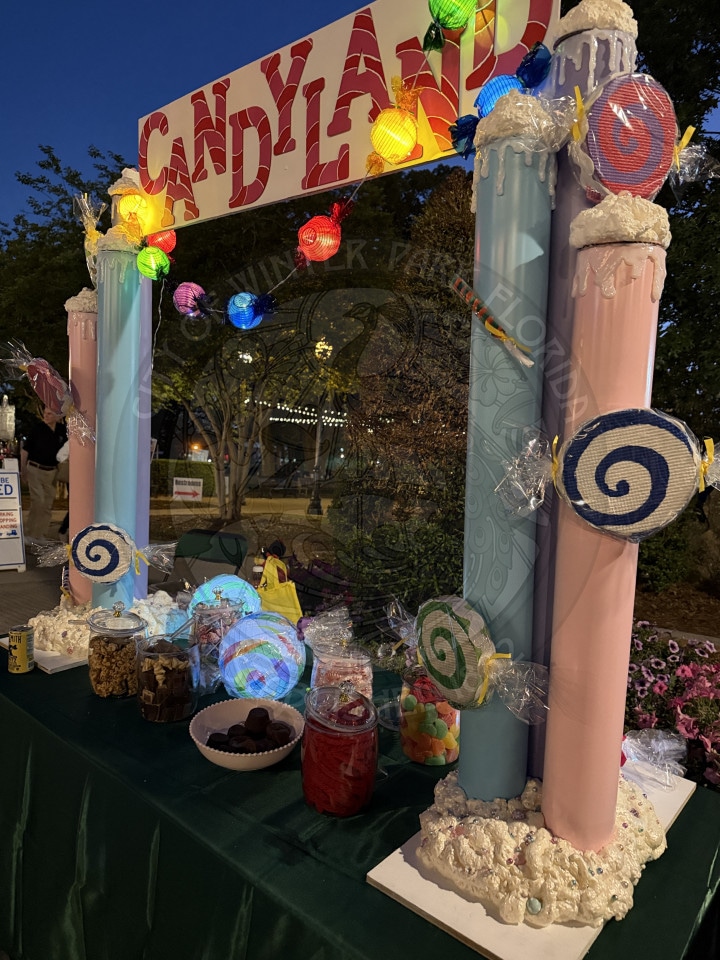 A colorful Candyland-themed booth with giant candy decorations and jars of assorted sweets on a green tablecloth.