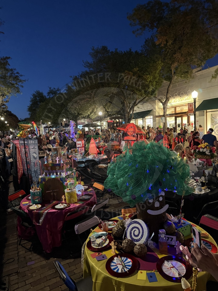 Colorful outdoor event at night with decorated tables, lights, crowds, and festive atmosphere under trees and shop lights.
