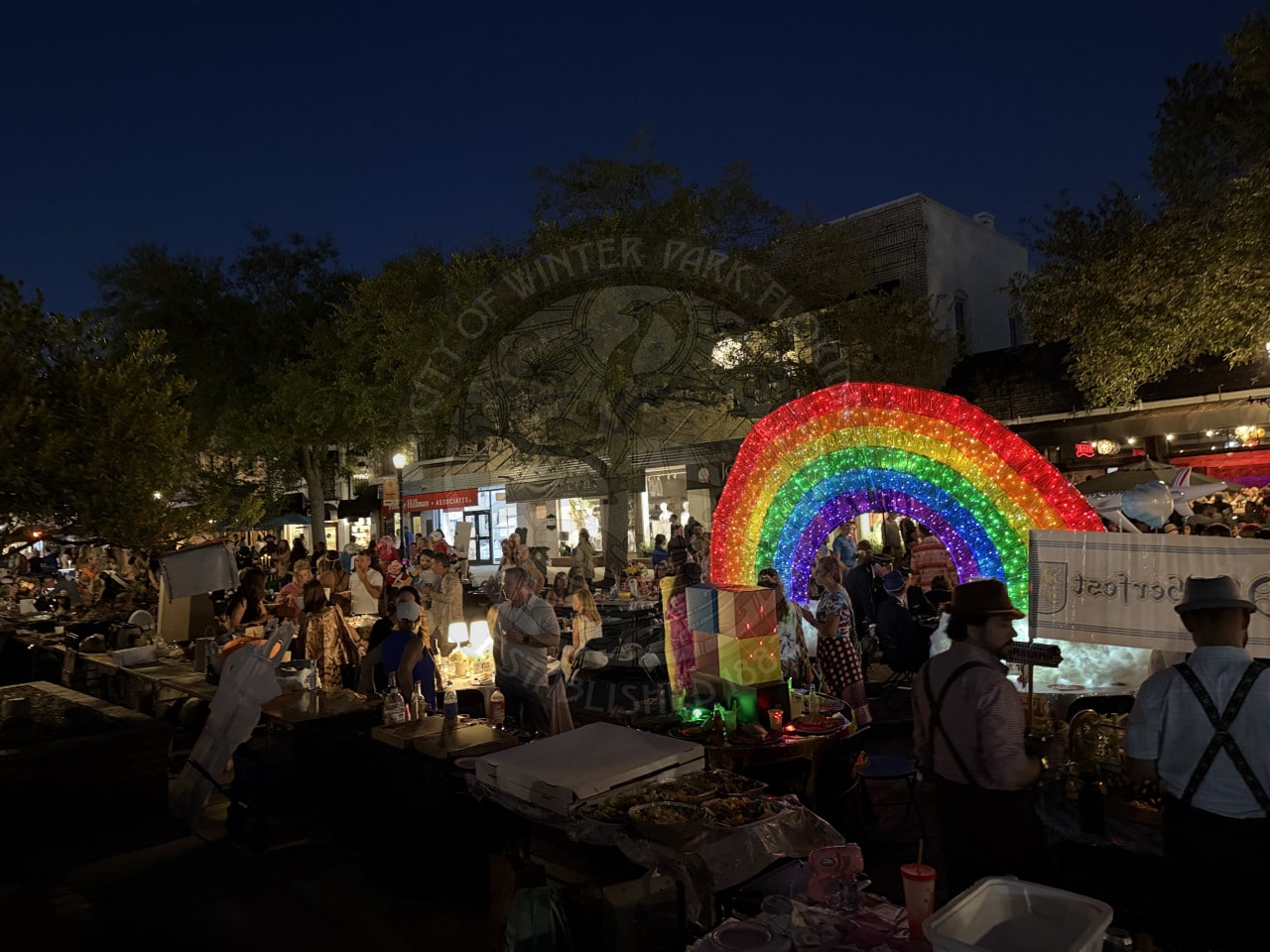 Outdoor night market with a crowd, food stalls, and a large illuminated rainbow decoration in the center.