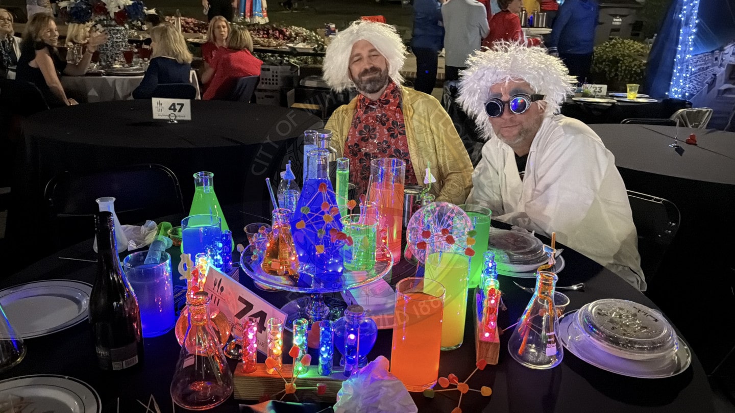 Two people in white wigs and costumes sit at a table decorated with glowing, colorful science lab props at a party.