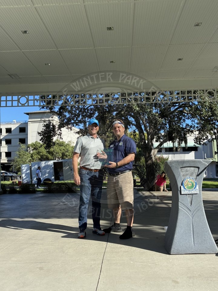 Two men standing outdoors, one holding a glass trophy.