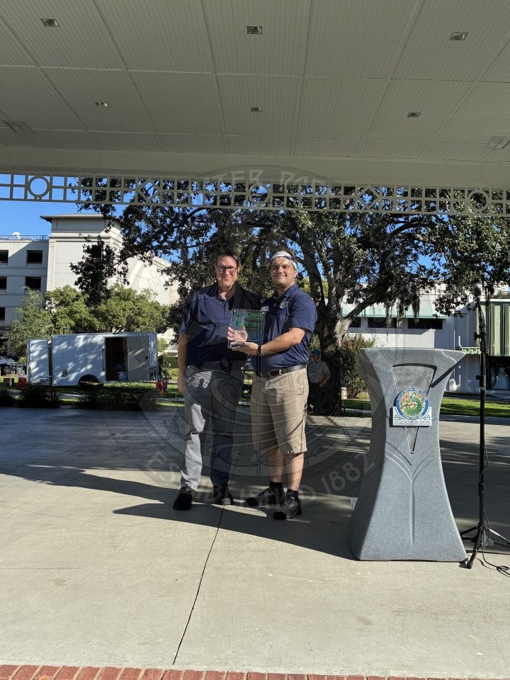 Two men stand outdoors under a pavilion, one presenting the other with a glass award.