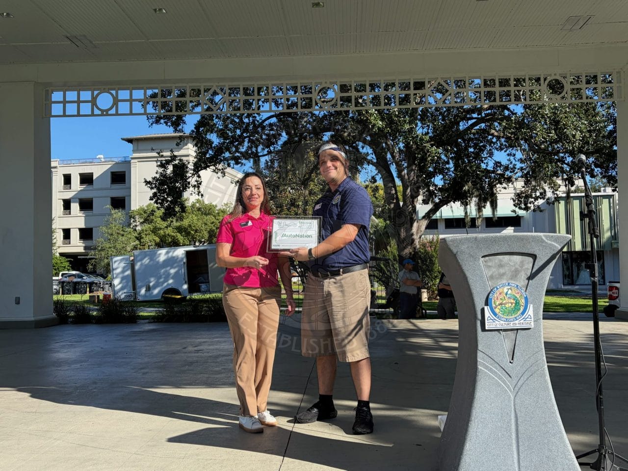 A woman in a pink shirt accepts an award certificate by a man in a blue polo shirt under a park pavilion.