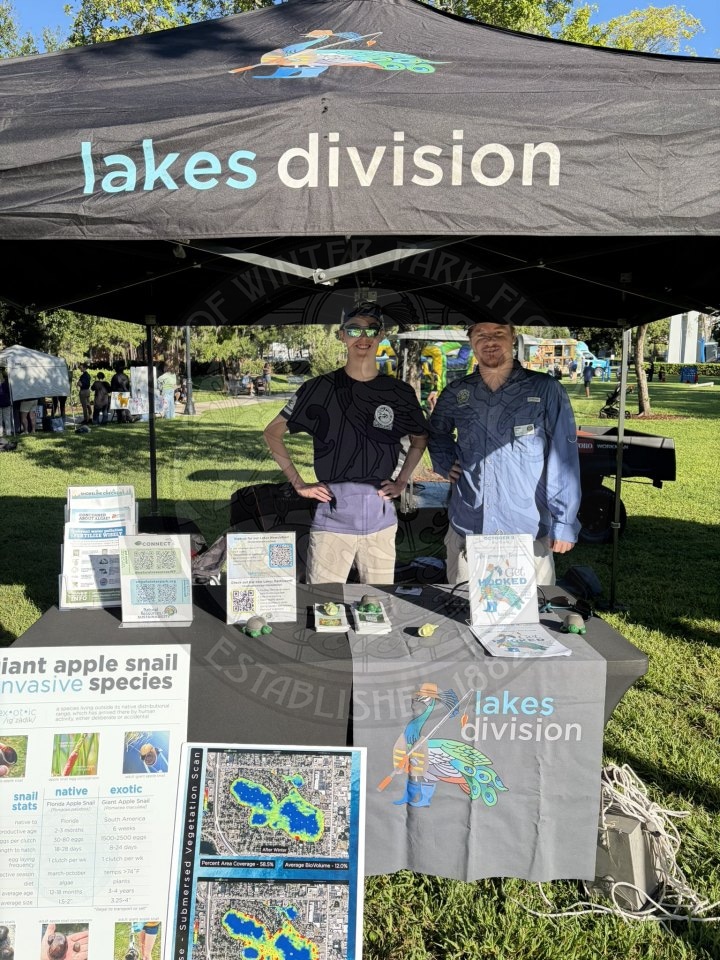 Two men stand behind a Lakes Division information booth at an outdoor event.