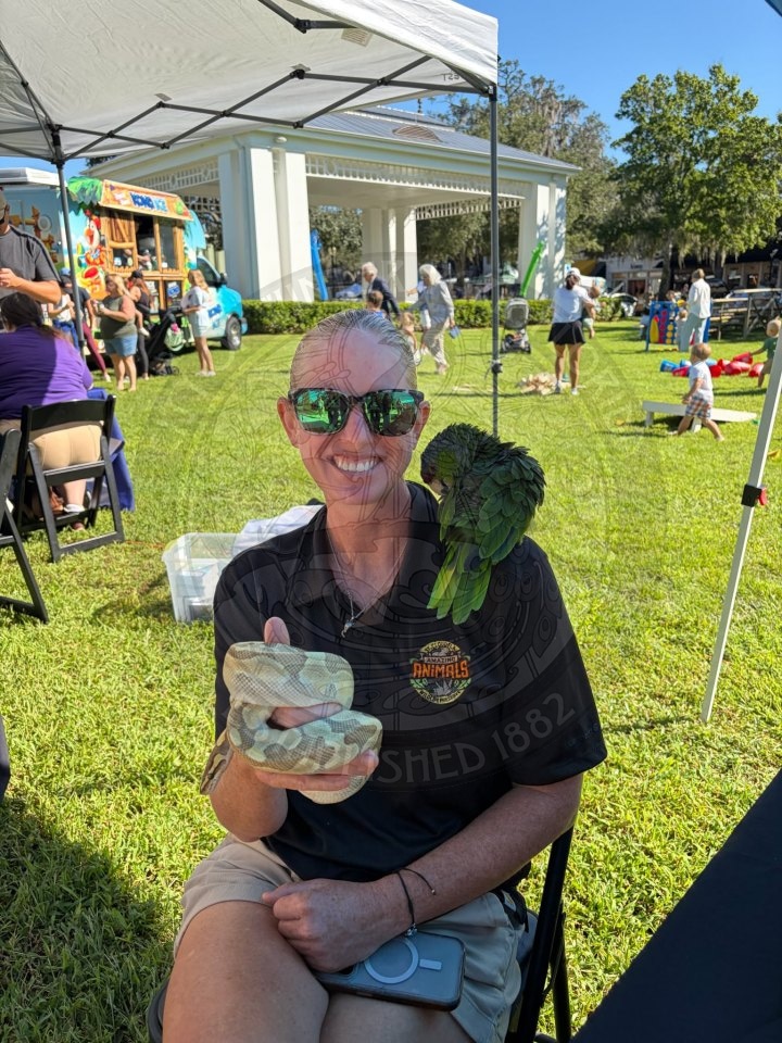 A smiling woman in sunglasses holds a snake and has a parrot perched on her shoulder at an outdoor park event.