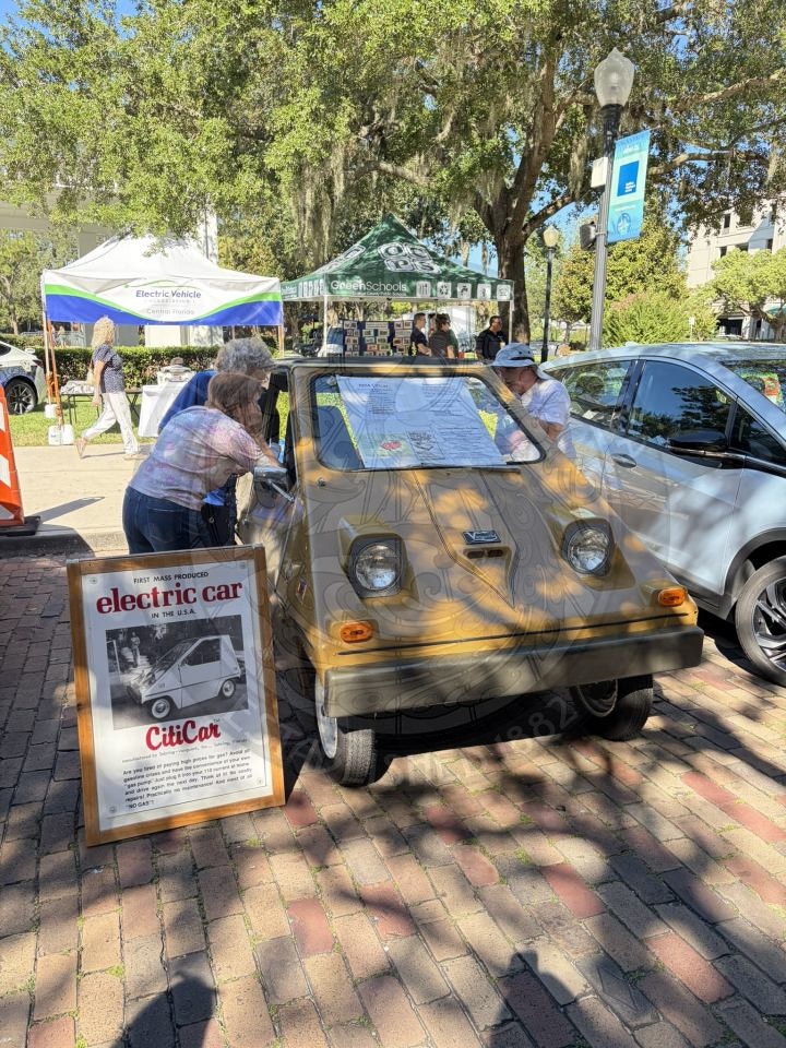People looking at a vintage tan CitiCar at an outdoor event with an informational poster standing in front of it.