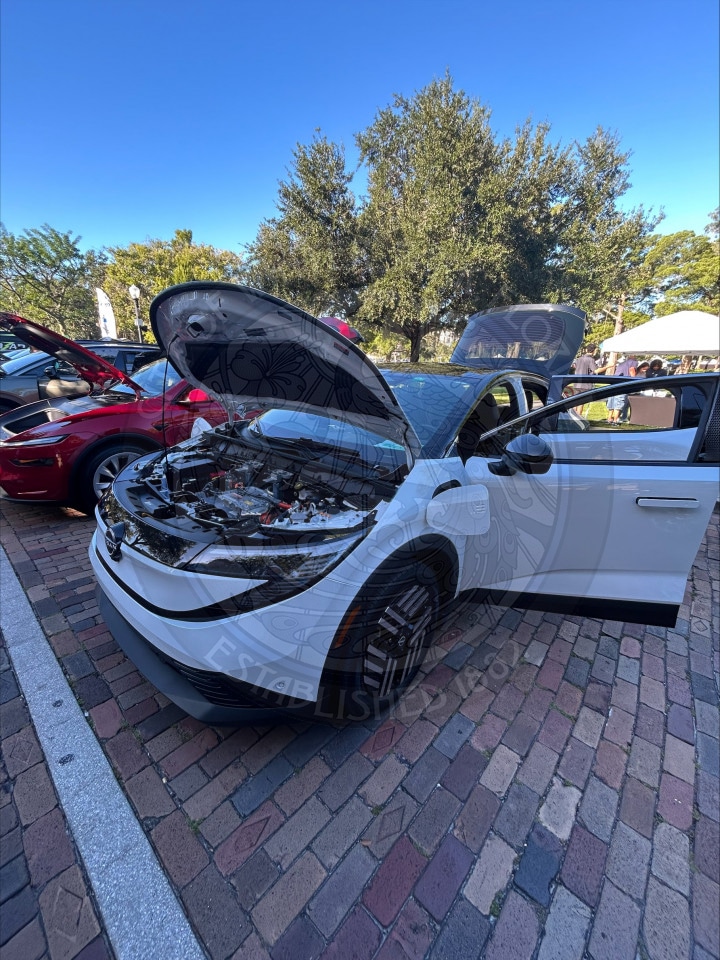 A white electric car with its hood and driver door open at an outdoor vehicle display.