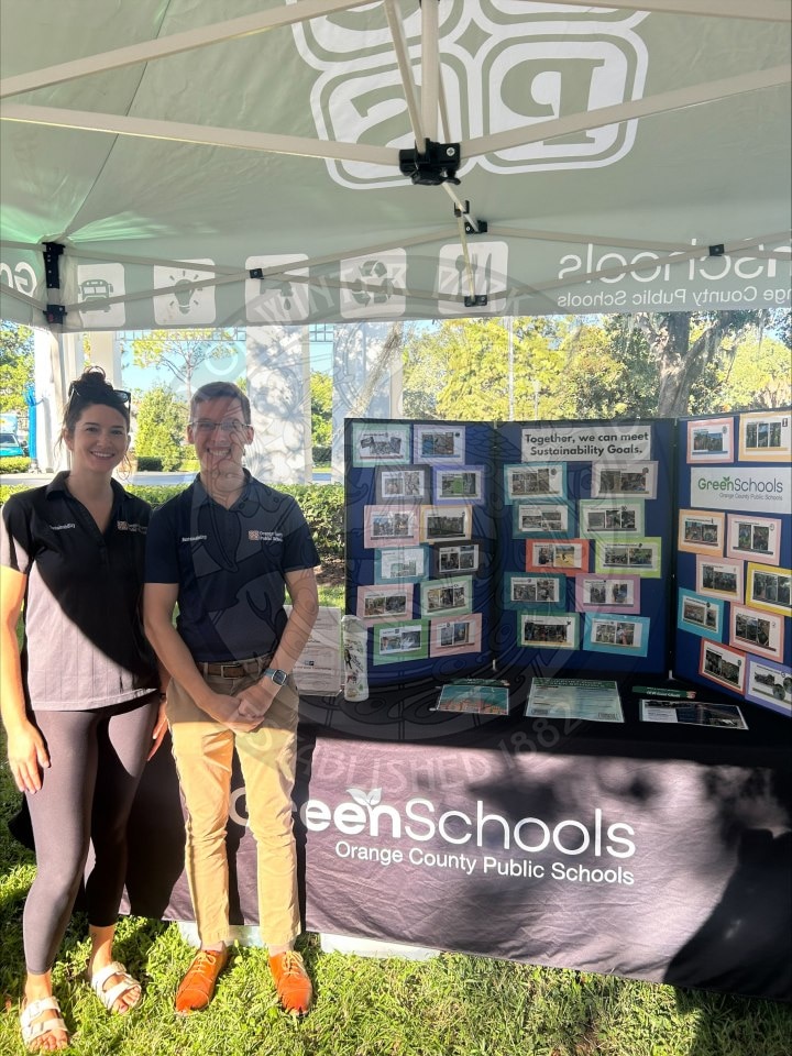 Two people stand in front of a GreenSchools display table under a canopy.