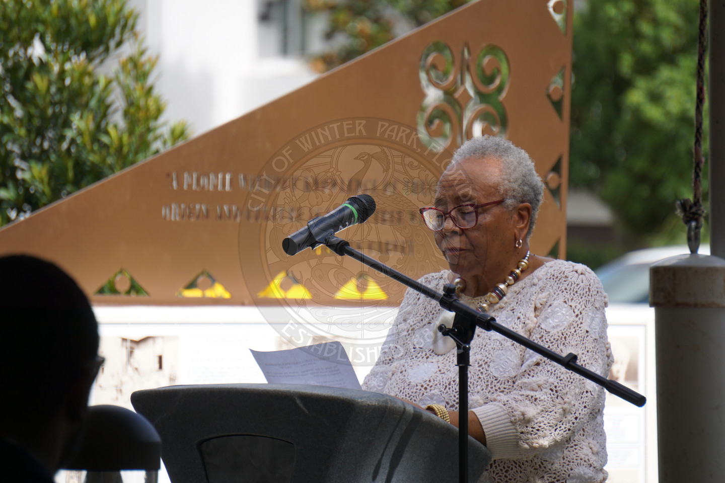 a woman standing behind a podium with the microphone pointed to her face while she is talking and looking at a paper.