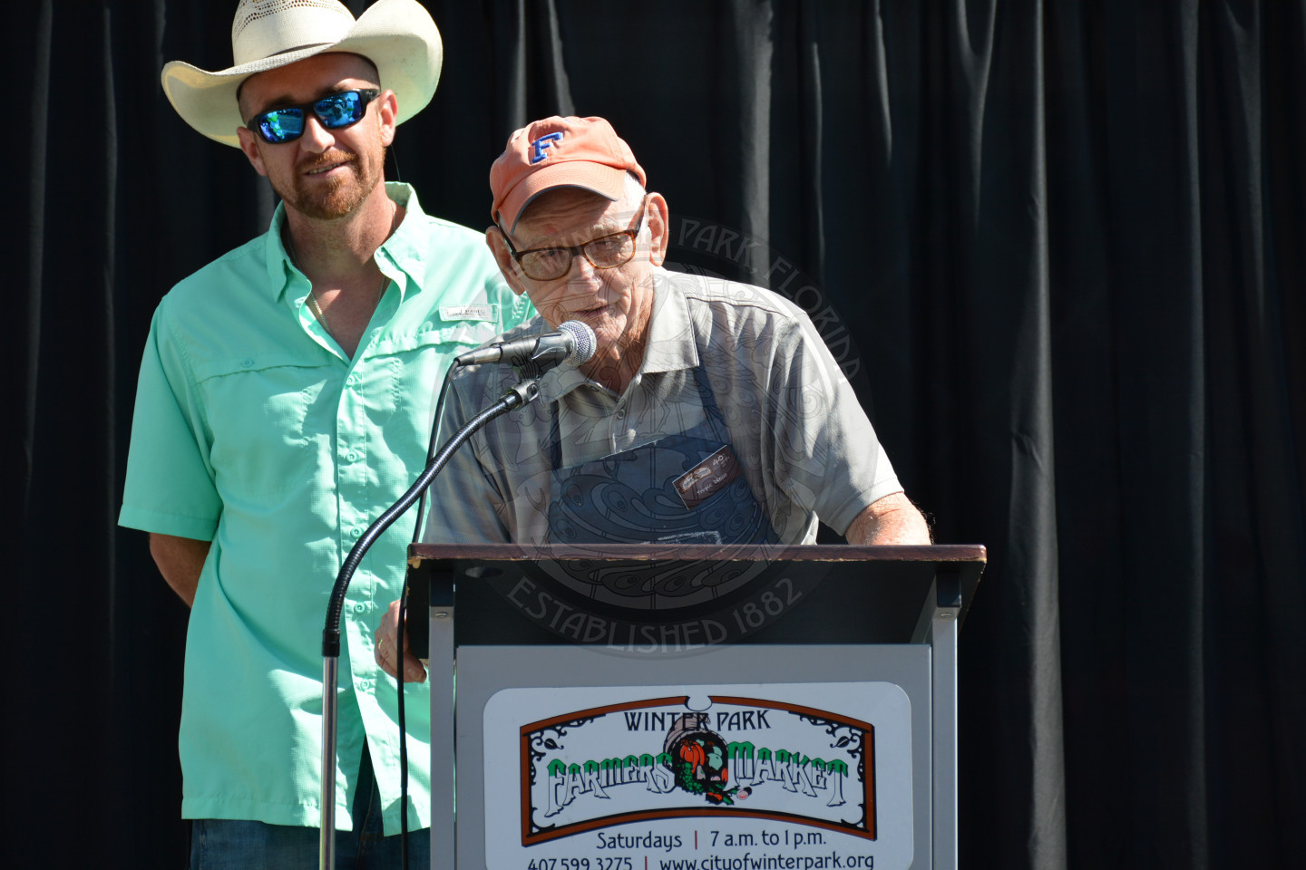 Gentleman with a light colored baseball cap, leaning to speak at the microphone on the podium on stage.  There is another gentleman standing behind him in a straw hat and sunglasses.