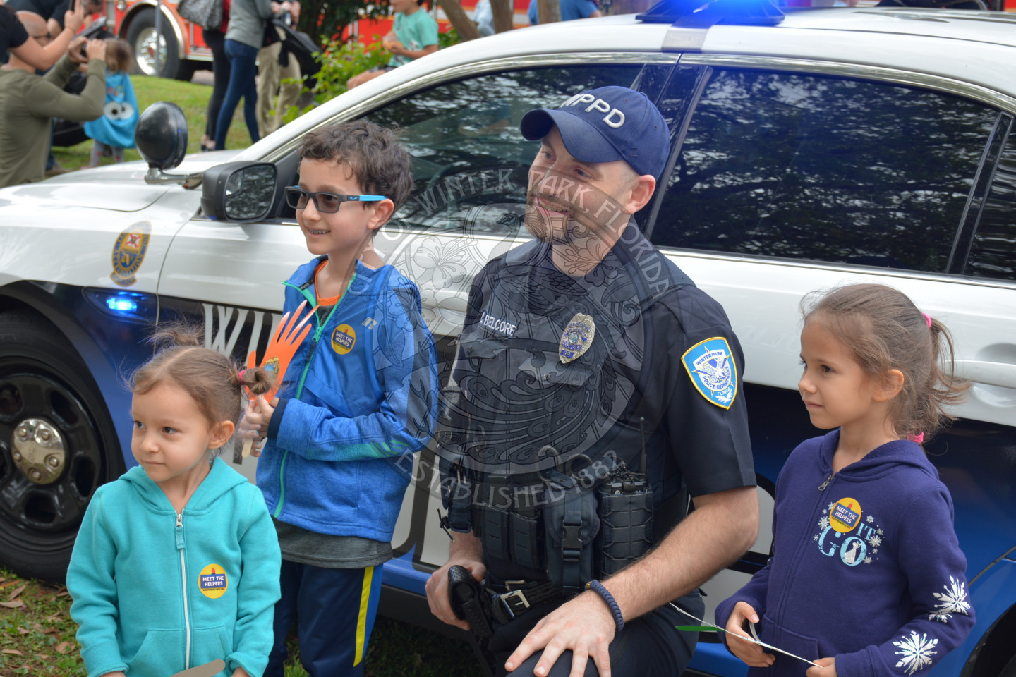 three children posing with police officer in front of police car