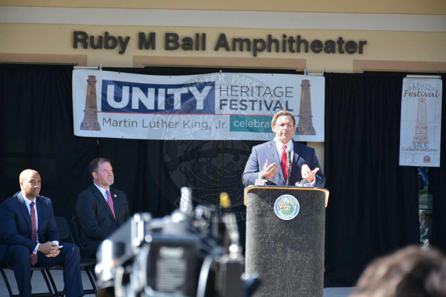 Governor Ron DeSantis speaking at the podium with the Unity Heritage Festival banner displayed behind him