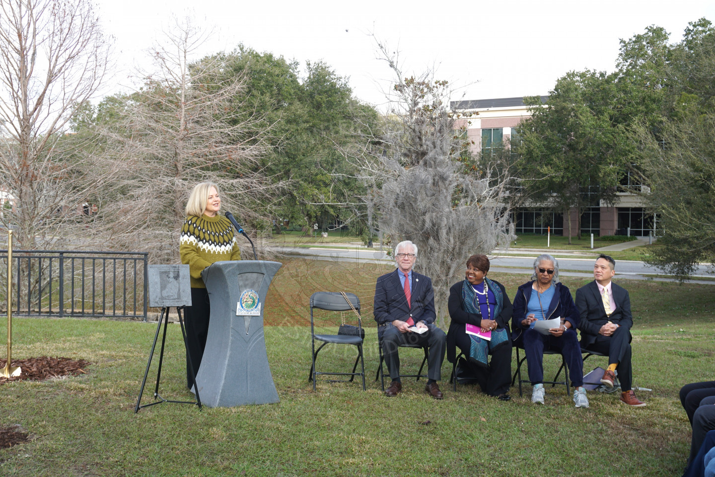 The assistant city manager standing behind a podium speaking into a microphone. To the right of her is five folding chairs with a group of four pepole sitting in them.