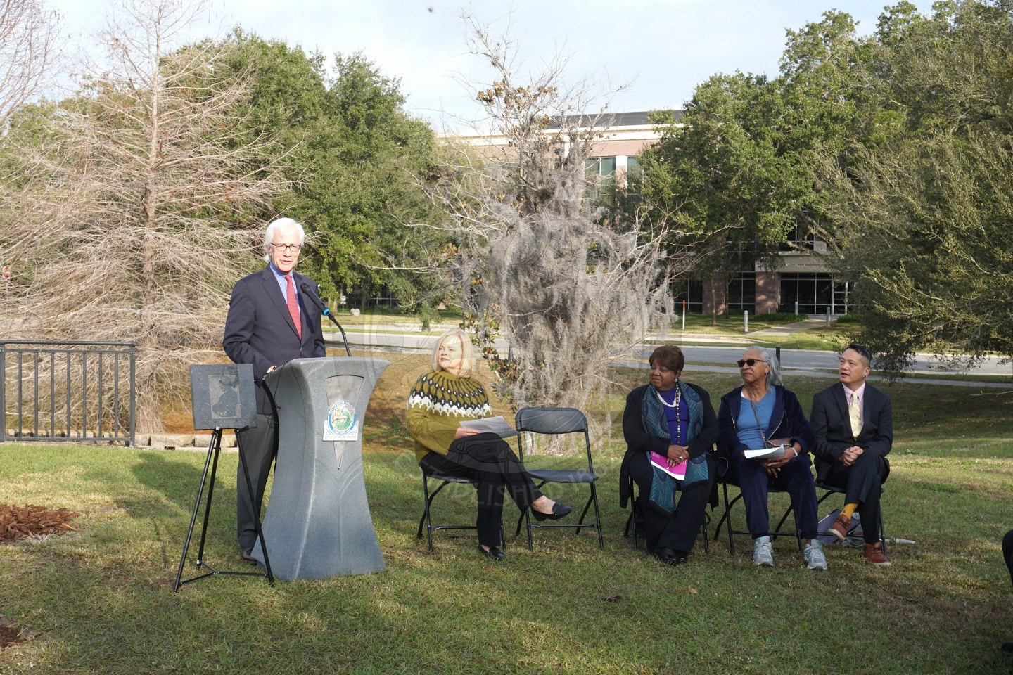 The mayor standing behind a podium talking to people who are sitting in black folding chairs in front of him. To the right of him is 5 black folding chairs with  4 people sitting in them.