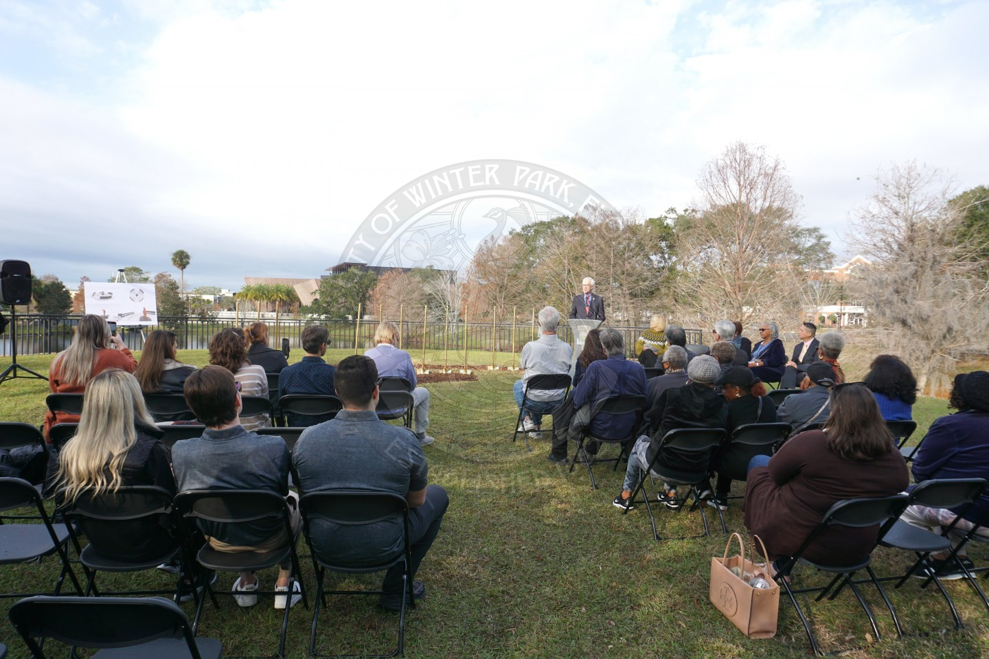 People sitting in black folding charis on the grass and facing the podium where the mayor is talking.