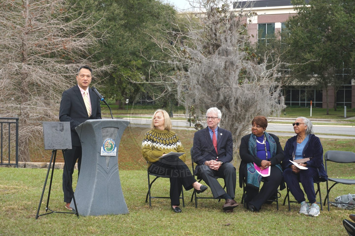 a man standing behind a podium speaking into the microphone to the audience whoa re sitting in front of him in black folding chairs. To the right of the podium is five black folding chairs with people sitting in four of the chairs.
