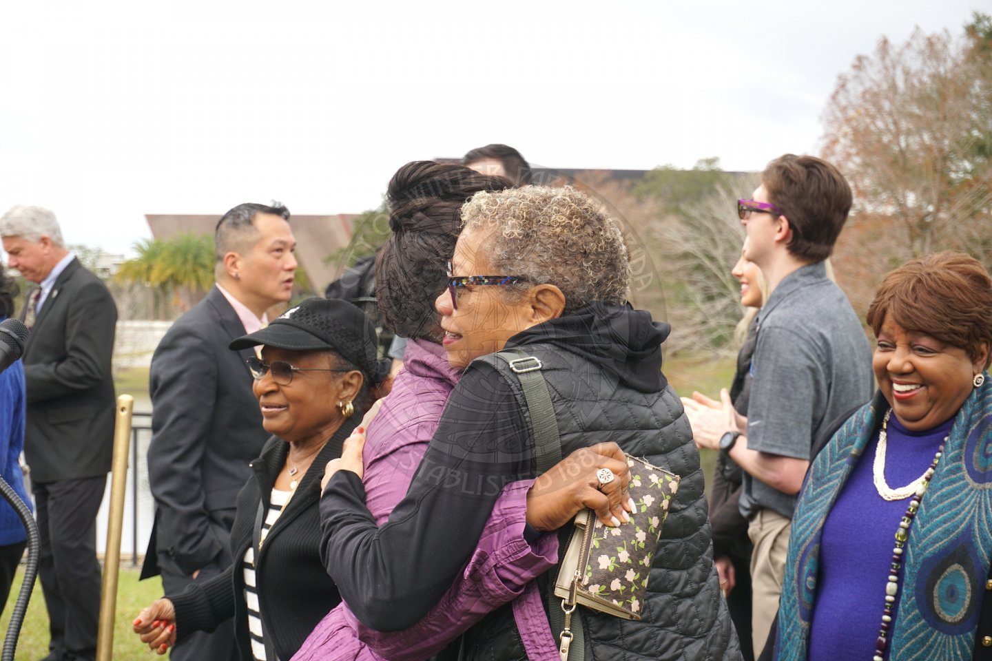 two ladies hugging with people mingling behind them.