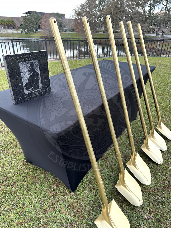 a table with a black linen and a statue that says Martin Luther King, Jr. Park and a picture of him. In front of the table is six gold shovels that are leaning against the table.