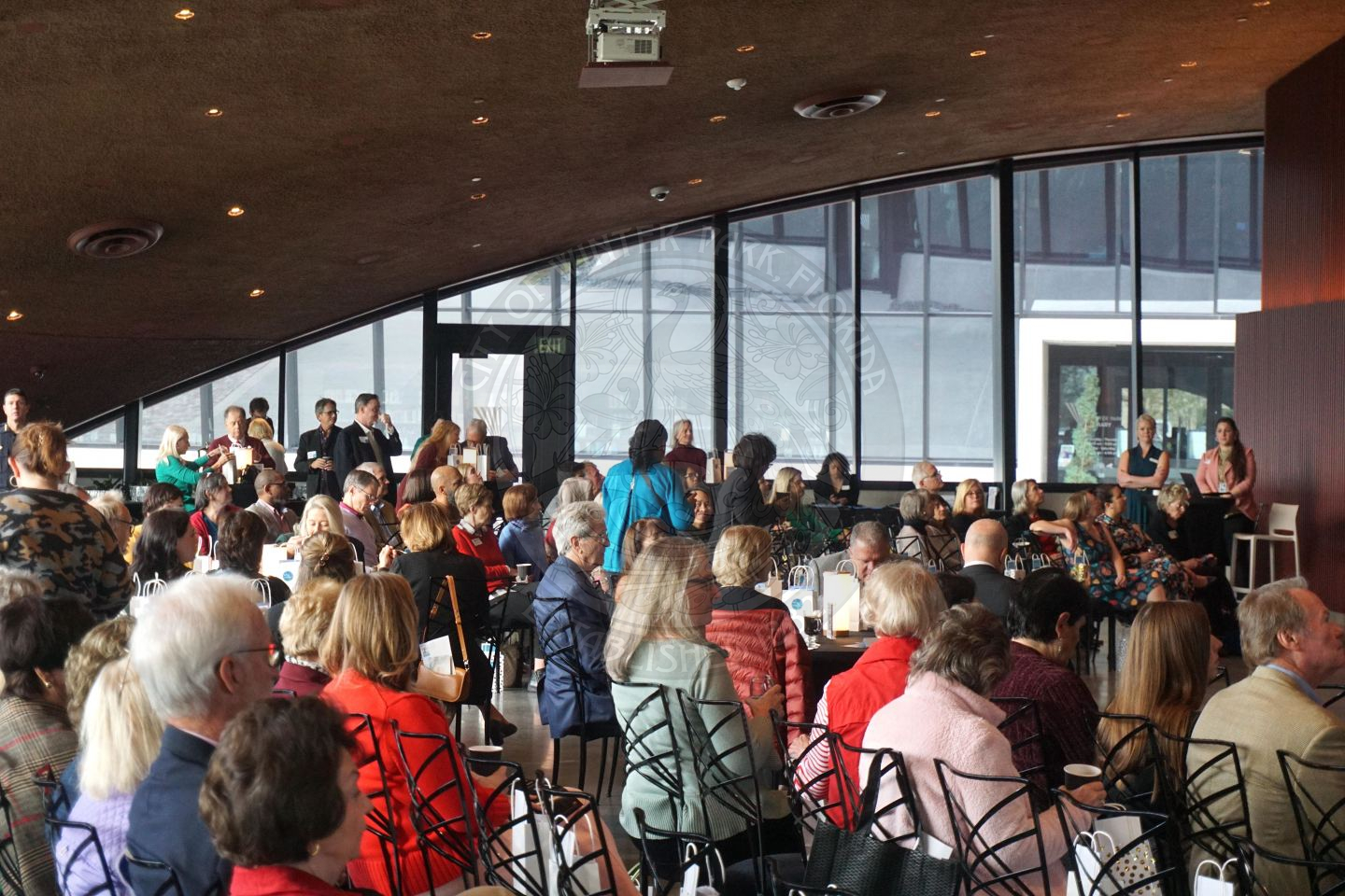 People sitting in chairs and tables and in the back of the venue standing by the hightops waiting for the event to start.