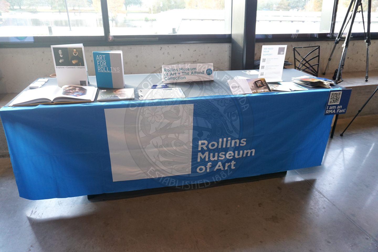 a table with blue tablecloth that says Rollins Museum of Art. On top of the table is information about Rollins Museum of Art (RMA) and pamplets to take home.