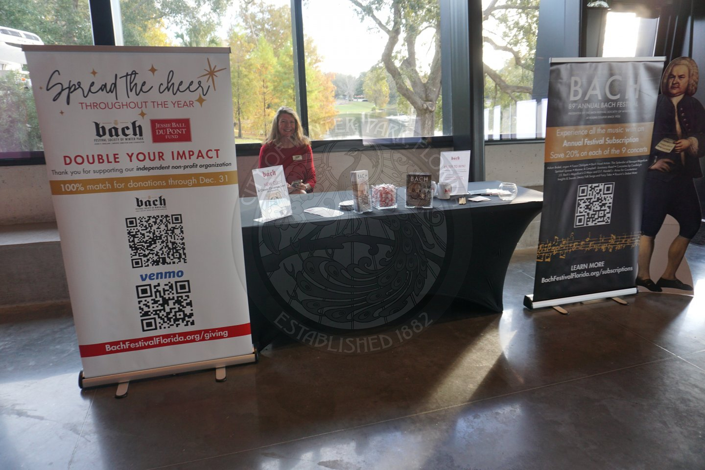 An 8 foot table with black linen and pamplets on the table giving information about Bach. There are 2 banners on each side of the table with QR codes to scan to get more information about the alliance member.