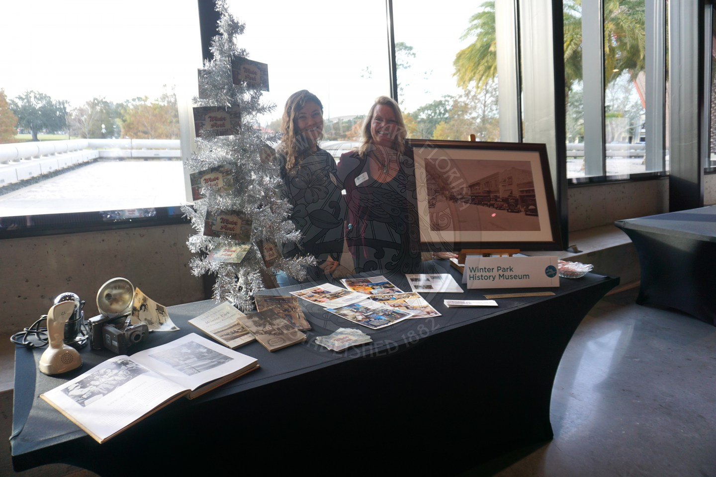 two women standing behind an 8 foot table with black linen on it. There is a christmas tree with photos in the tree. Around the Christmas tree there is a picture frame on easel with papers on teh table talking about Winter Park History Museum.