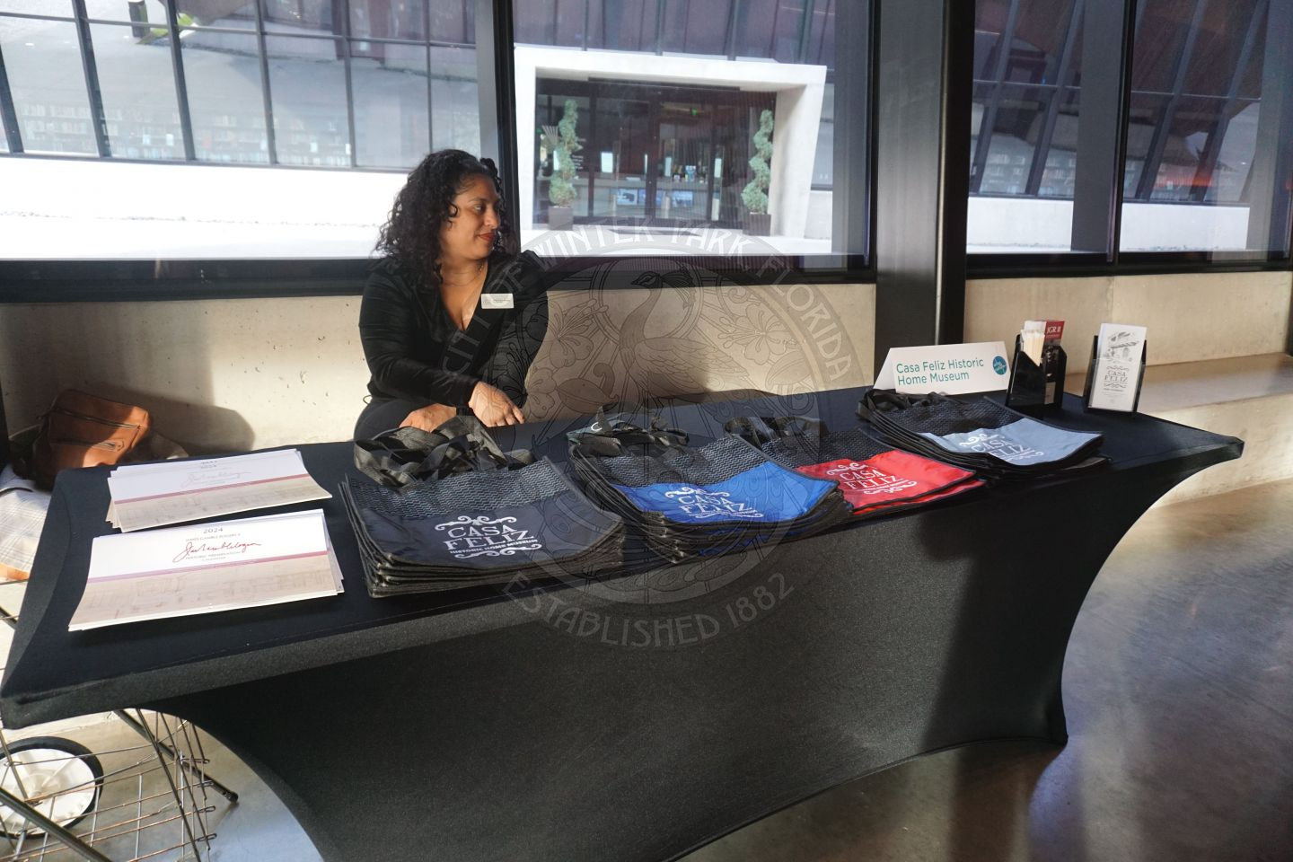 a table with black linen and a lady sitting behind the table. There are tote bags that say Casa Feliz on them and information next to the tote bags about the venue.