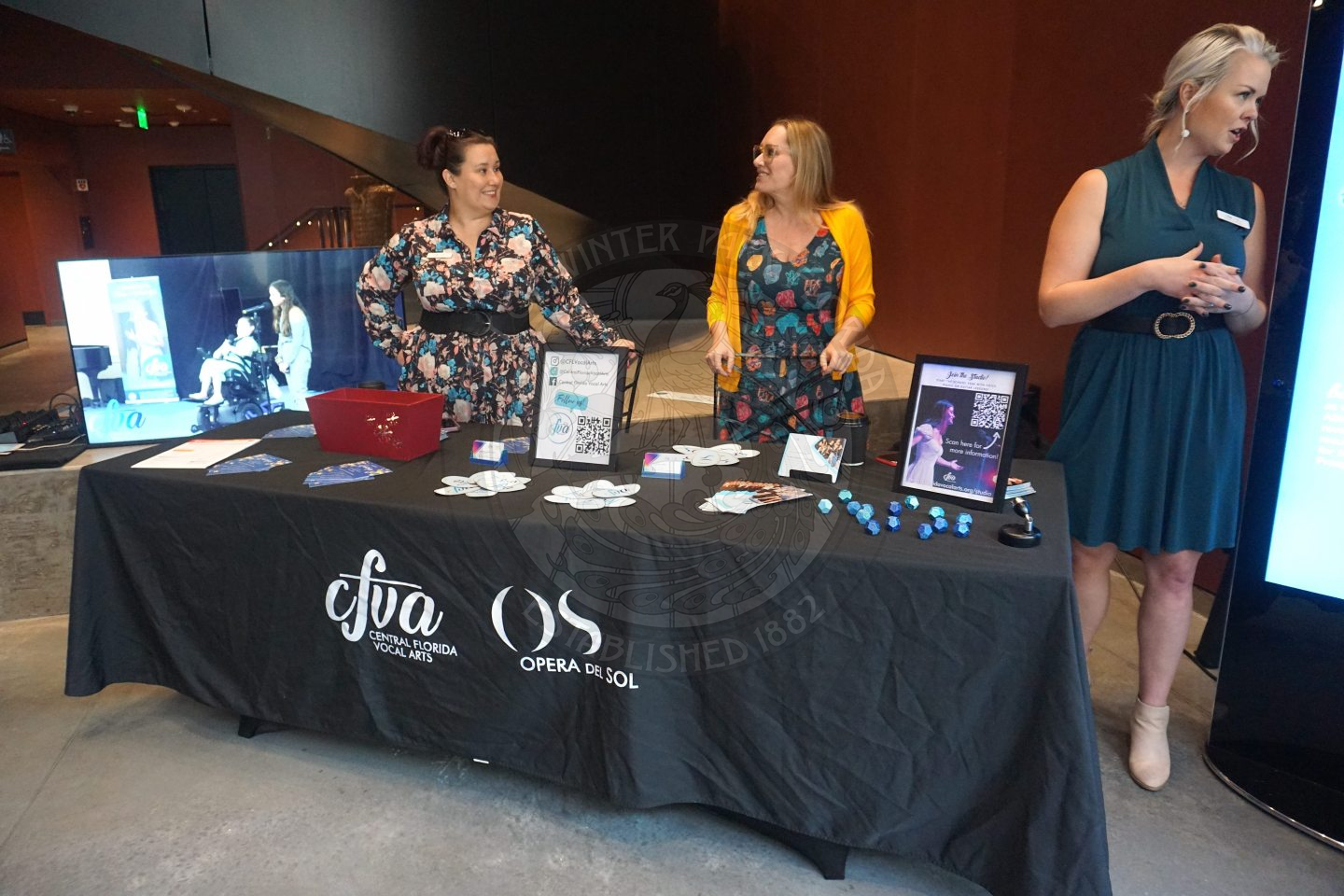 A table with a black linen that says "CFVA Central Florida Vocal Arts". There is a small tv screen to the left and on the rest of the table there is information 