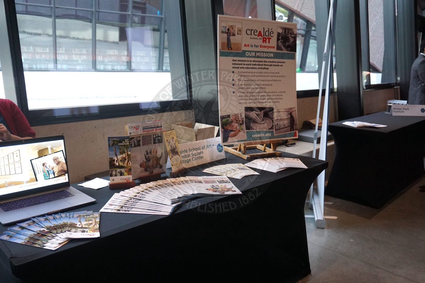 A table with a laptop on it and pamaplets to take home talking about Crealde and Hannibal Sqaure Heritage Center. on the right side of the table there is an easel with a poster board filled with information on Crealde School of Art.