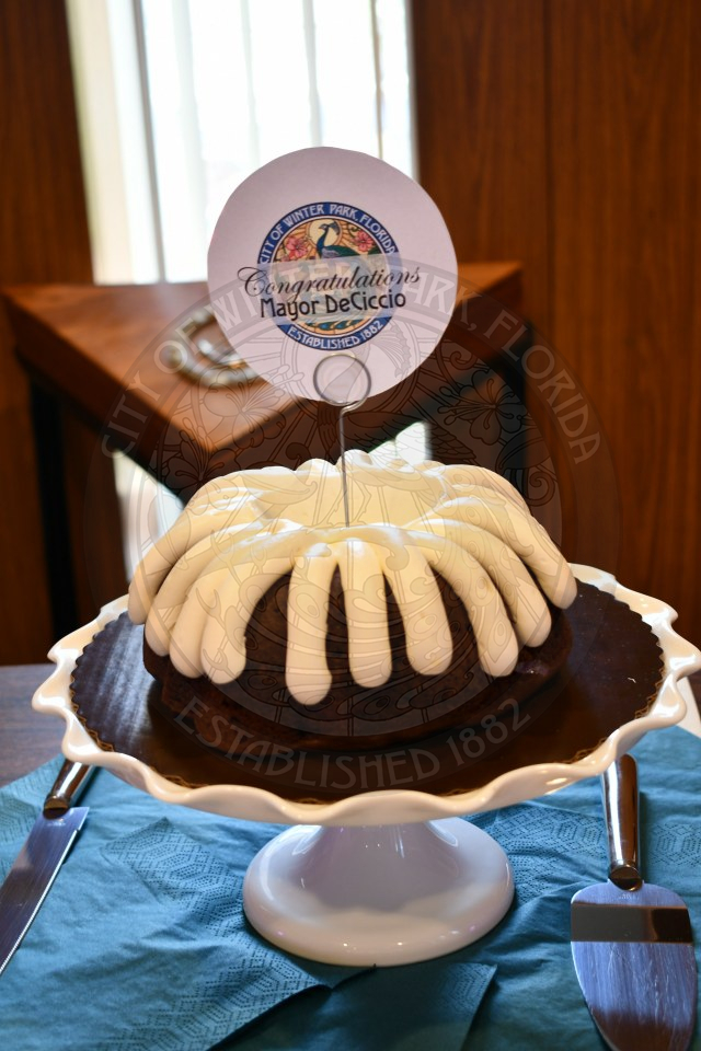 Chocolate bundt cake with white cream cheese frosting on the top. In the middle of the cake is a circular sign that says Congratulations Mayor DeCiccio.