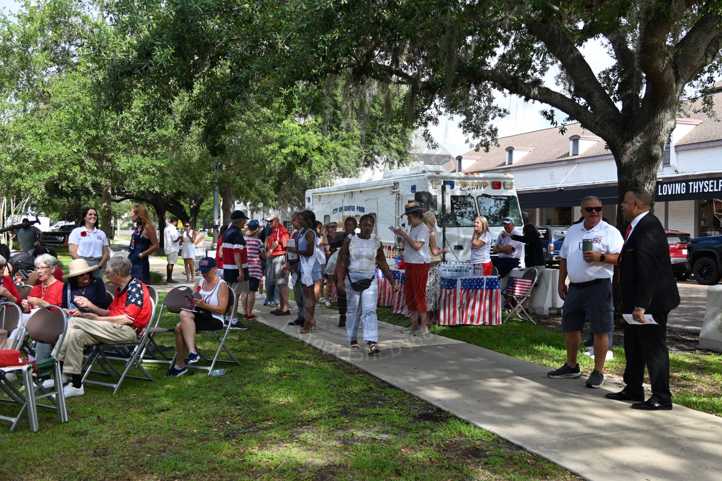 People gather outdoors near a command center truck, many wearing patriotic clothing, surrounded by trees and grass.