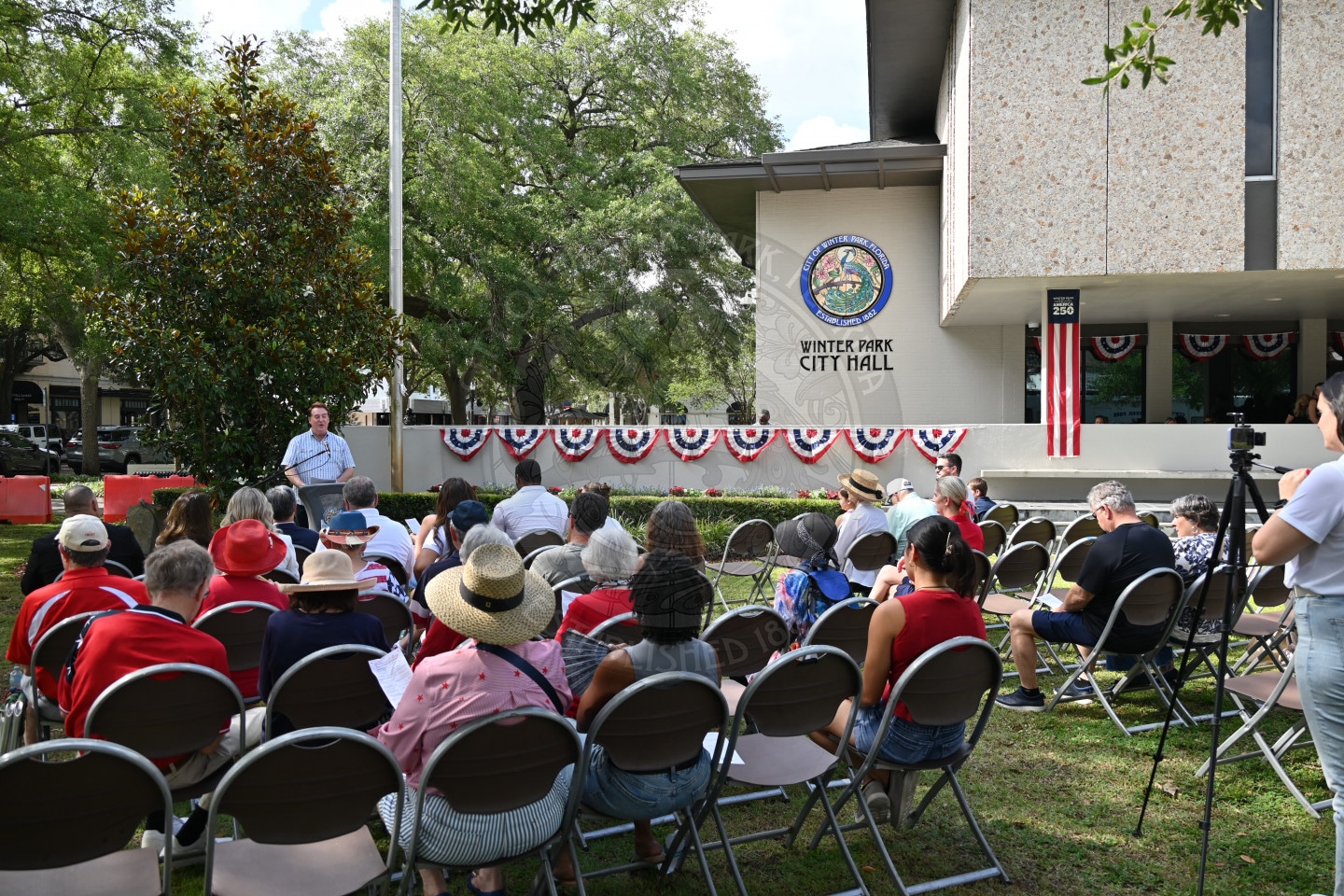 People seated on lawn chairs at an outdoor event in front of Winter Park City Hall decorated with patriotic bunting.