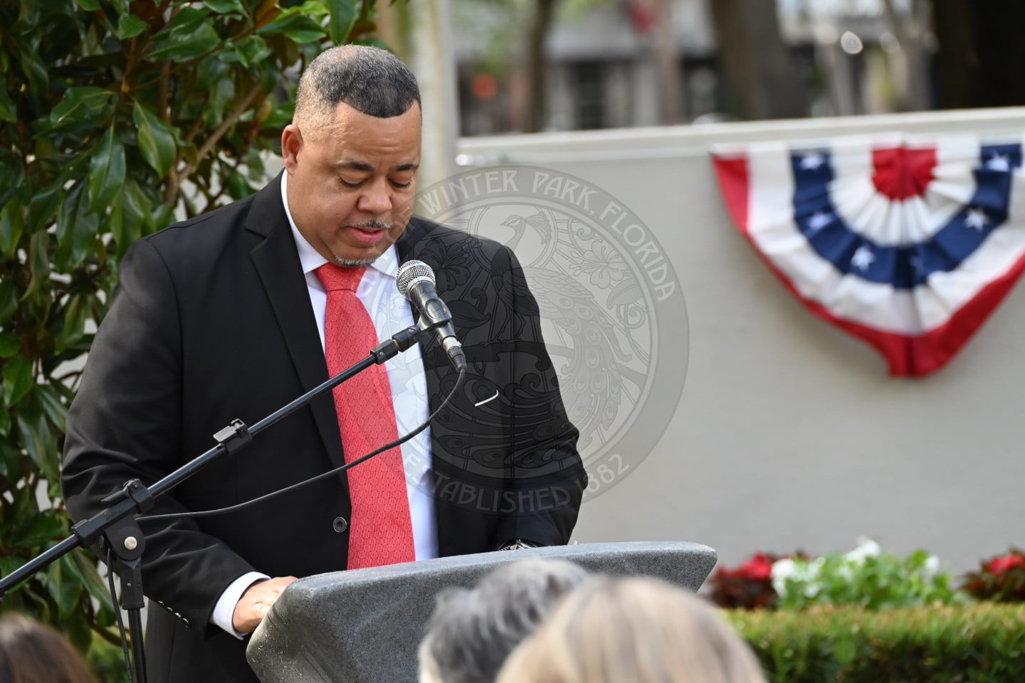 Man in a suit and red tie speaks at a podium outdoors, with patriotic bunting and greenery in the background.