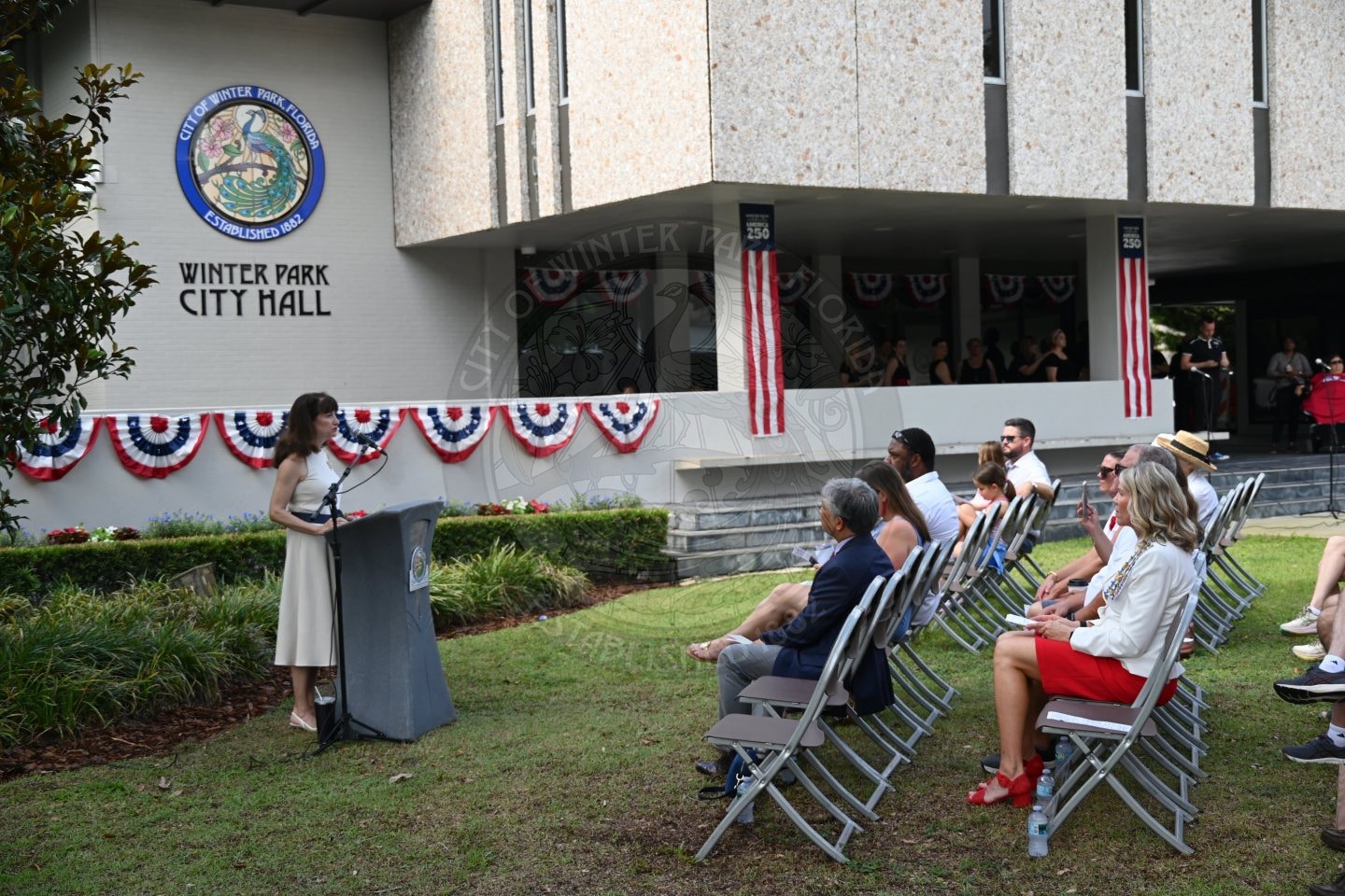 A woman speaks at a podium to seated people outside Winter Park City Hall decorated with patriotic bunting.