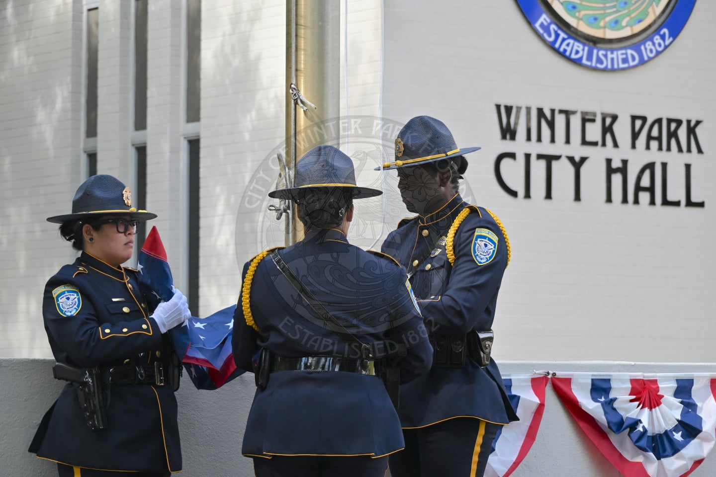 Three officers in uniform hold an American flag in front of a flag pole at Winter Park City Hall.